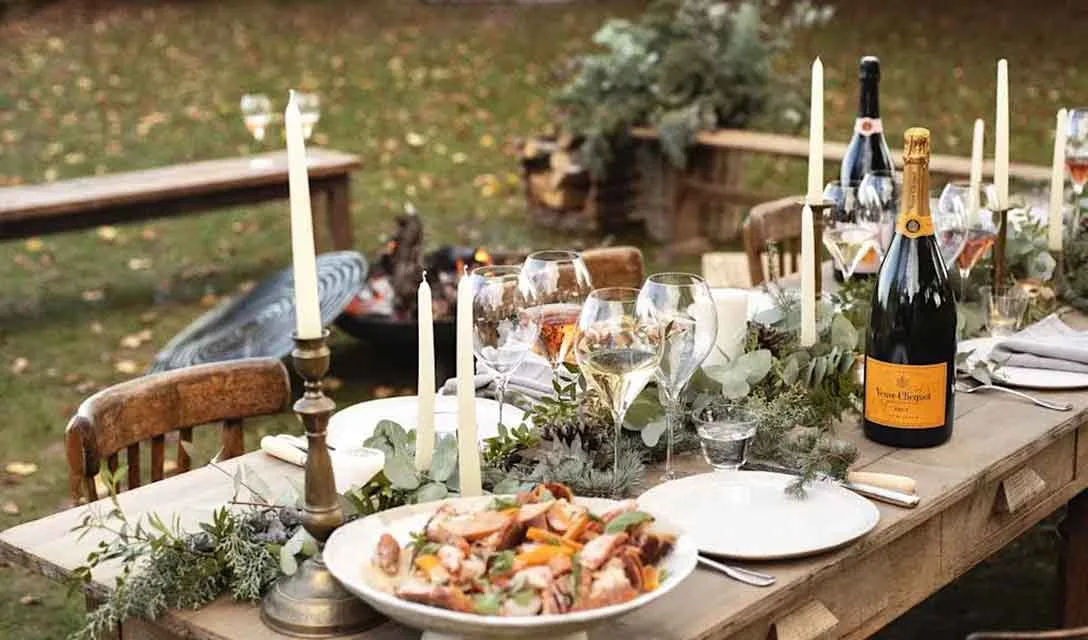 Outdoor table set with champagne, glasses, candles, and sharing dishes surrounded by autumn foliage.