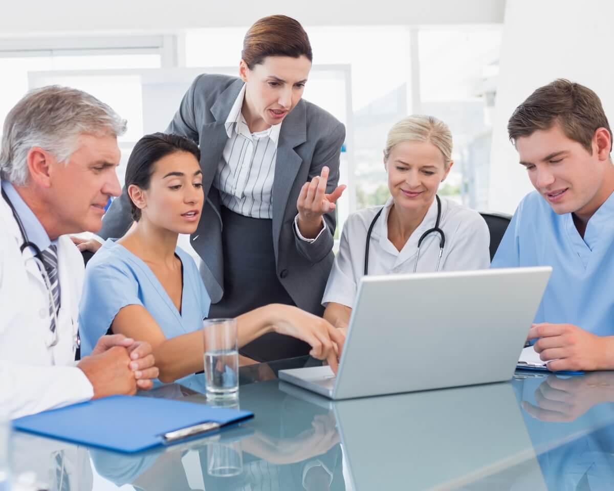 Group of health care professionals, dressed in suits, lab coats, and scrubs, review information on a laptop