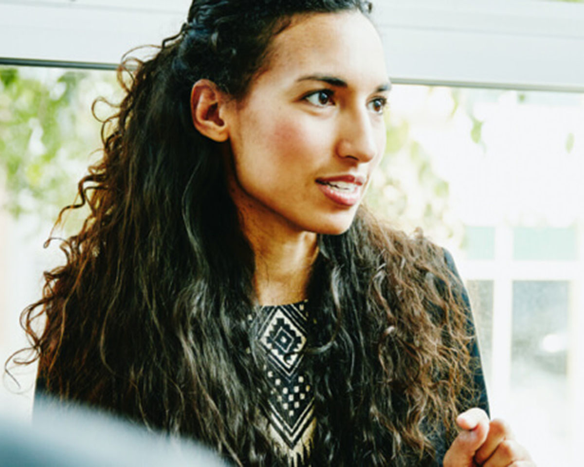 Professional woman with long curly hair wearing patterned black and white blouse engaged in conversation near bright windows