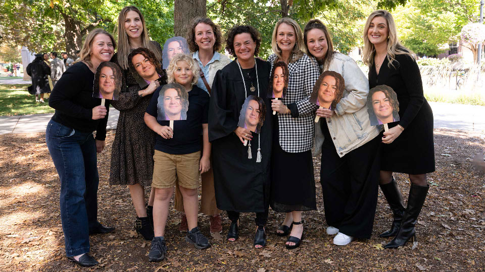 Leanna Chroman, wearing her black graduation gown and a white honors cord, stands outside with a group of her loved ones.