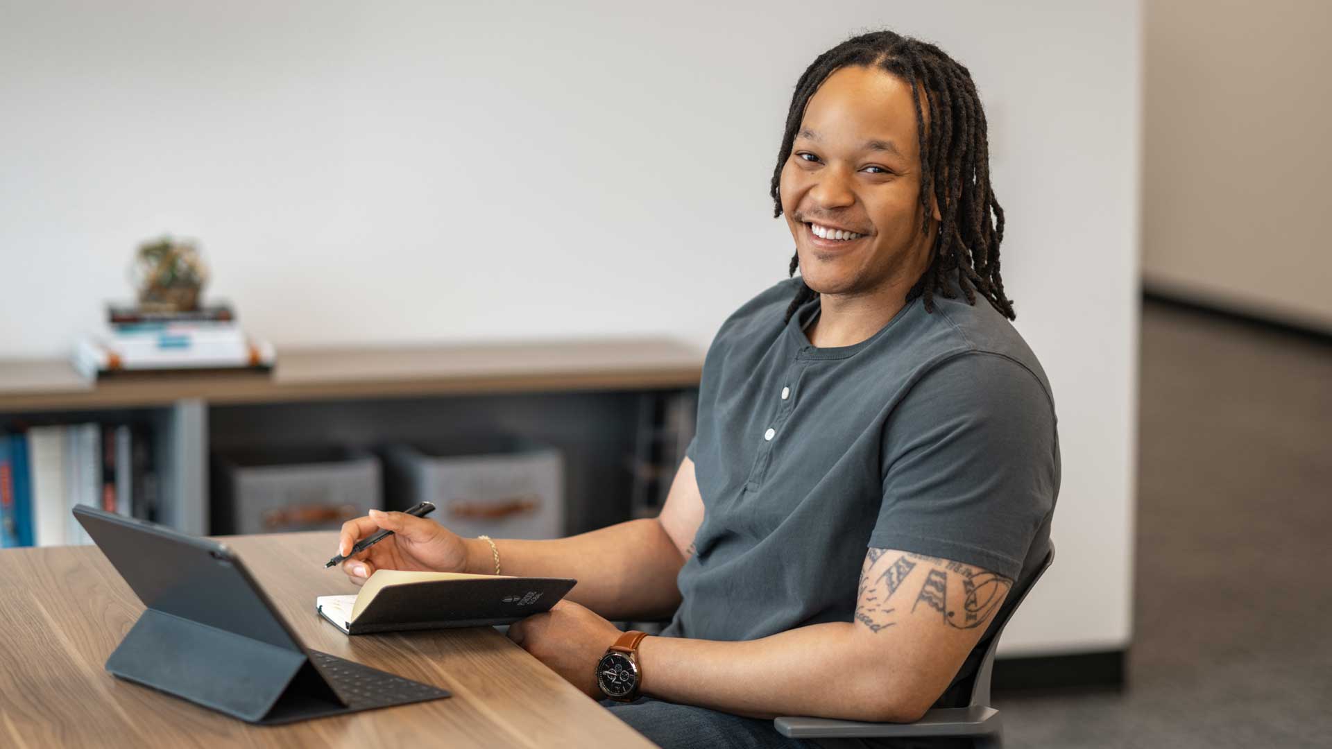 TJ sits at a table with his tablet and a notebook and pen, smiles at the camera.