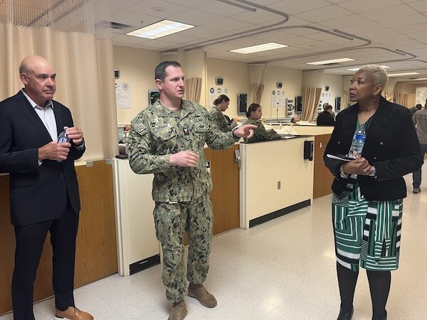 Two Purdue Global delegates meet with a man in military uniform in a METC facility. In the background are medical bays with students in military uniform attending to mannequin patients on beds.