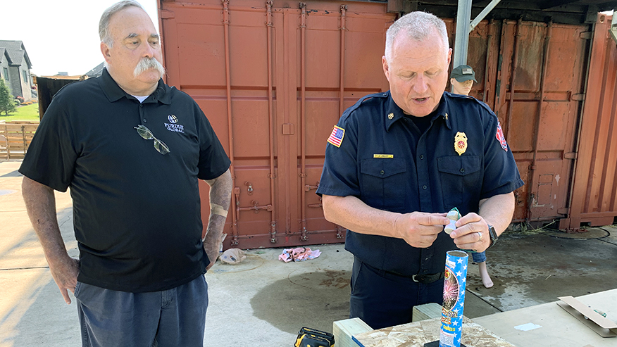 Two men stand in front of a table with fireworks.