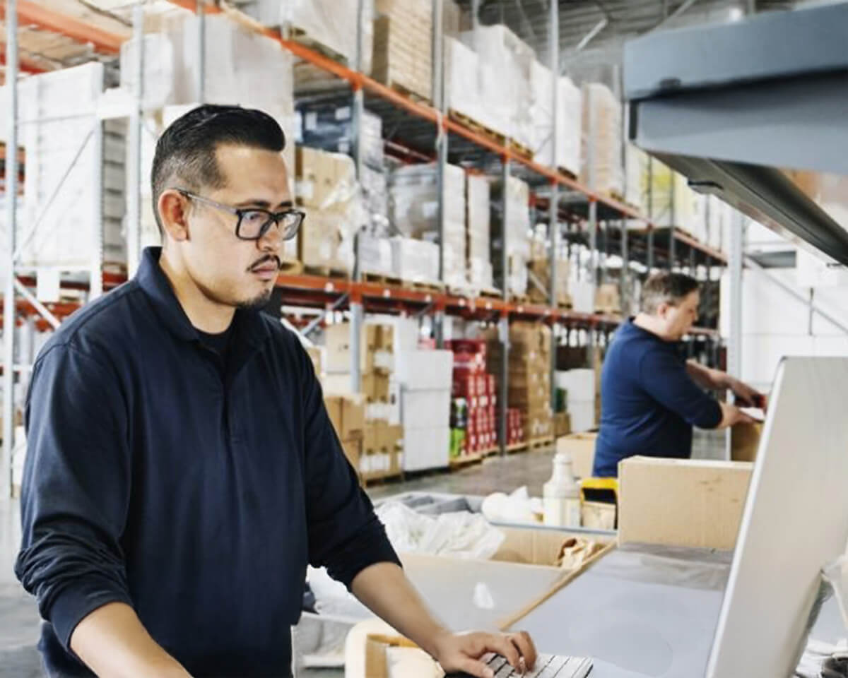 Warehouse worker in navy uniform and glasses using computer at workstation with storage shelves in background