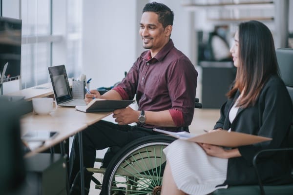 Two IT professionals, one in a wheelchair and one in an office chair, sit at a table and look at a laptop.