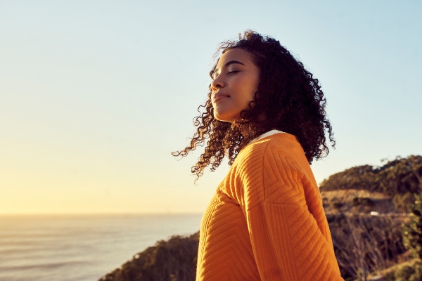 A woman with closed eyes stands near a coastline.