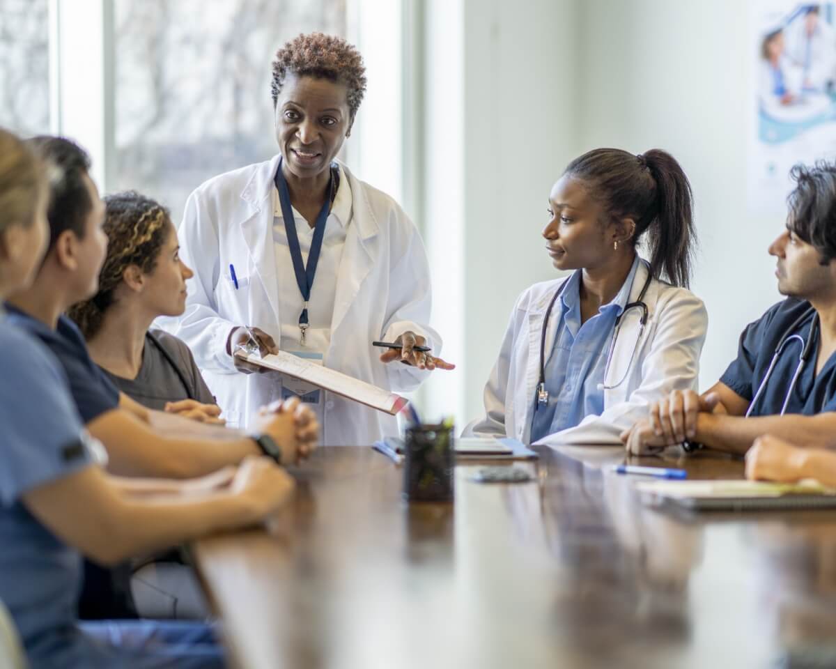 a nurse in a lab coat leads a meeting of nurses in lab coats and scrubs