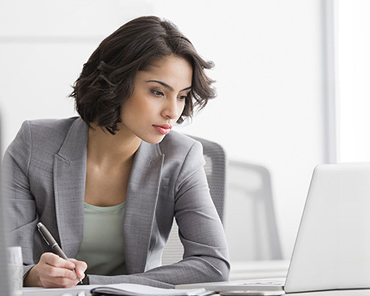 Professional in grey blazer with short dark hair writing at desk in bright office setting
