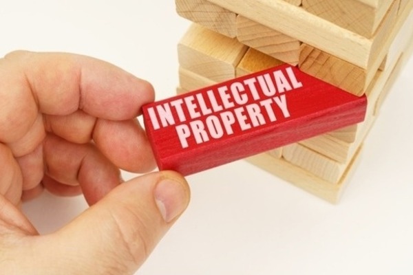 A hand pulling a wooden block labeled "INTELLECTUAL PROPERTY" from a Jenga-style tower of wooden blocks.