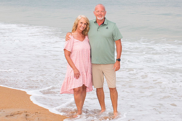 Sherry and her husband are smiling and ankle-deep in the water on a beach. She's wearing a pink sundress and he has a light green polo and tan shorts.
