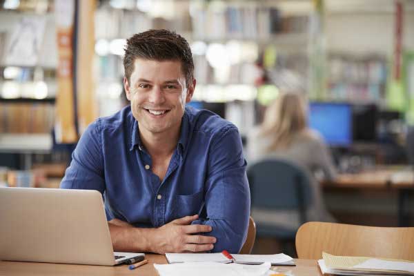 Smiling man in a blue dress shirt sitting in a library working on a laptop