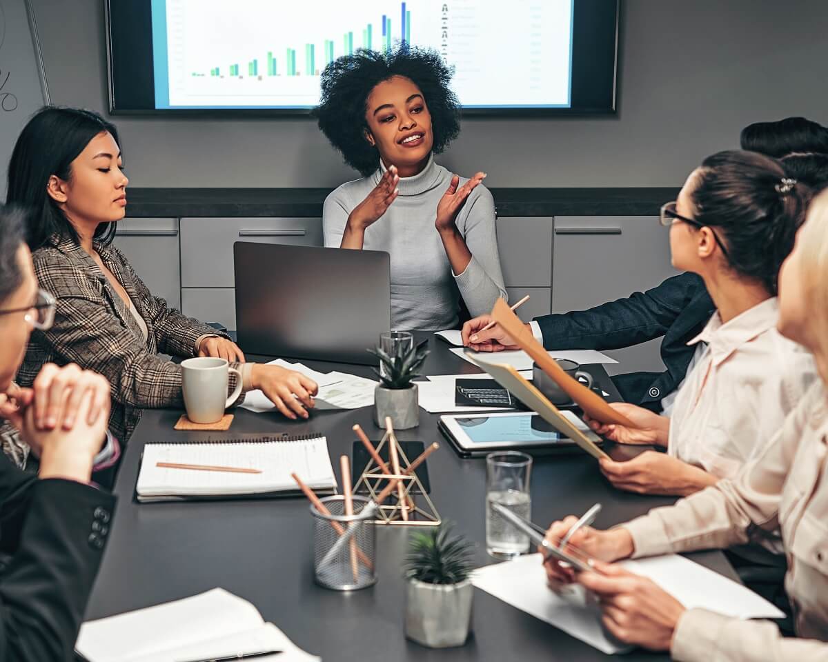 Professionals in meeting room take notes at a conference table and discuss bar graph presentation displayed on a smart screen
