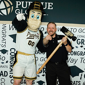 Purdue Global graduate next to the Boilermaker mascot, standing in front of a congratulations word cloud background