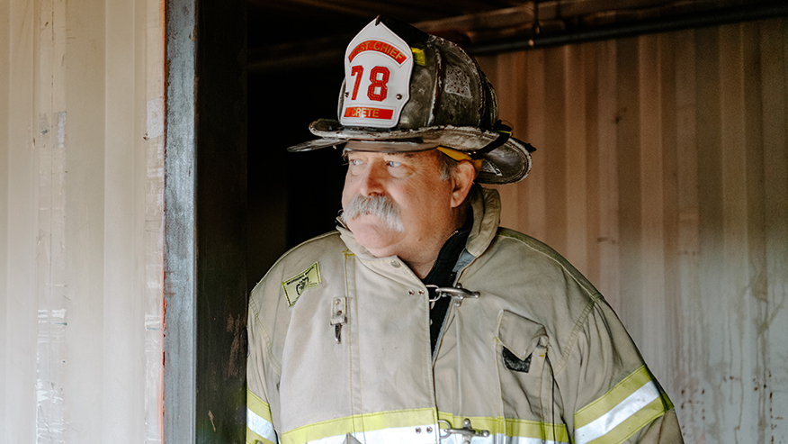 A firefighter with a heavy, white mustache wearing a coat and a fire helmet marked 78 looks into the distance.