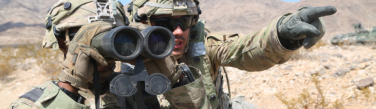 Soldier in camouflage gear using binoculars during field training in a desert mountain environment