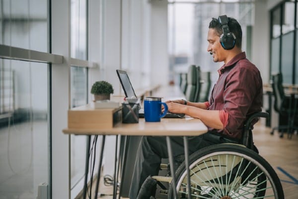 A computer support specialist sits at a desk with a laptop and talks to a computer user