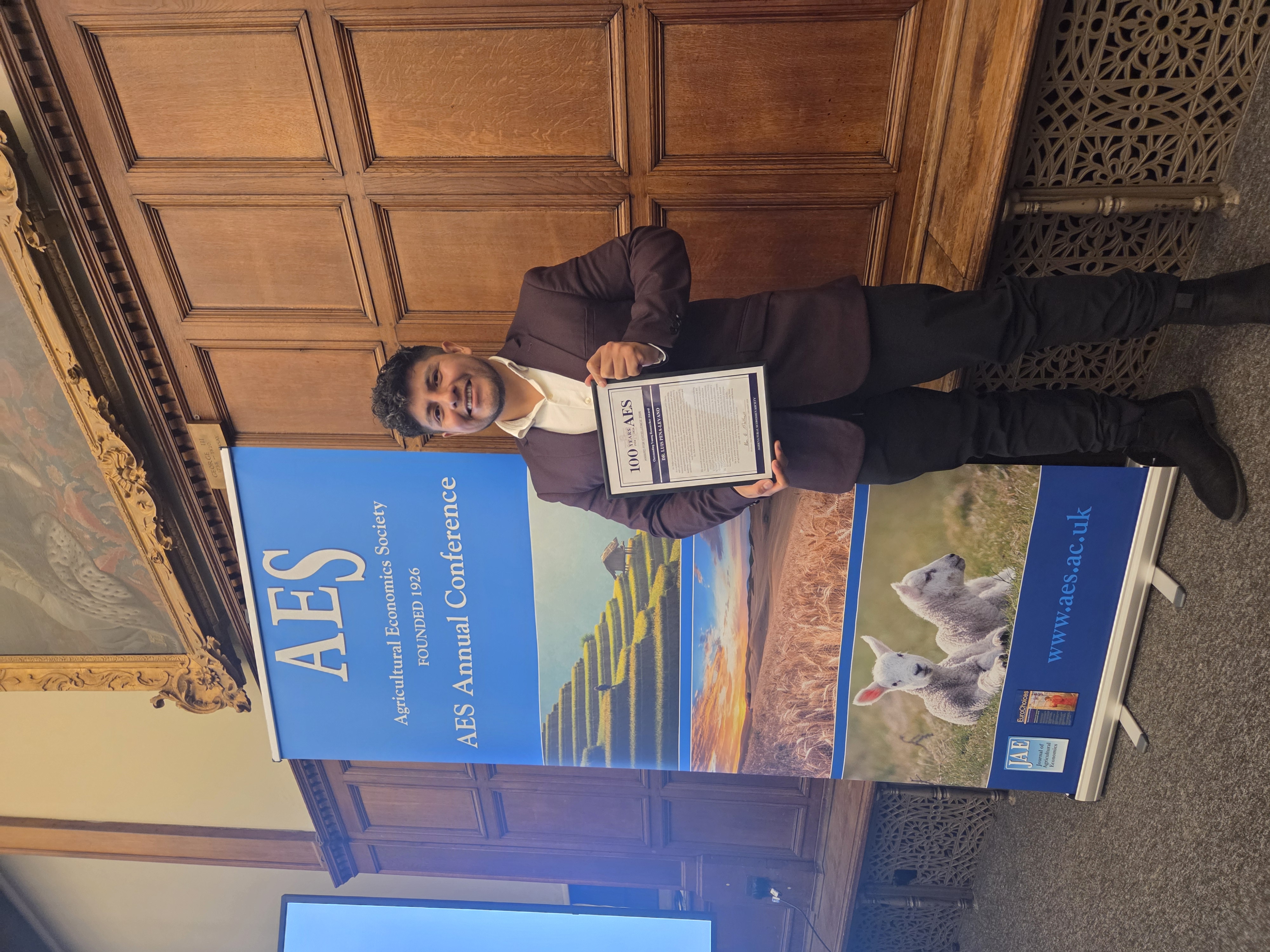 Cap: Purdue Global economics faculty member Luis Peña-Lévano was honored by the Agricultural Economics Society in March. (Photo provided)
Alt: A man in a suit with a plaque next to a blue banner. 