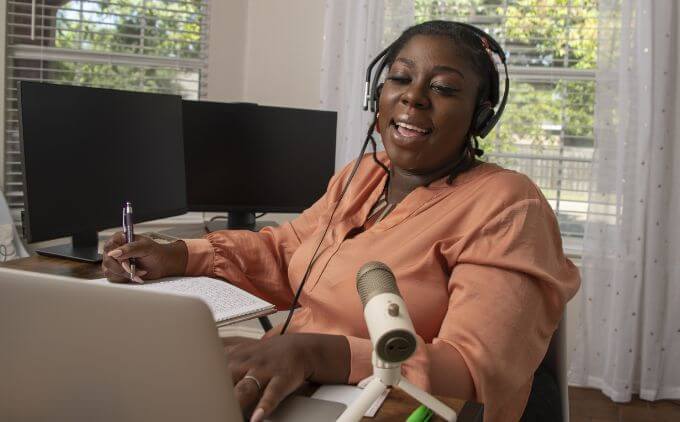 woman wearing a headset, taking notes, and working on a laptop at home