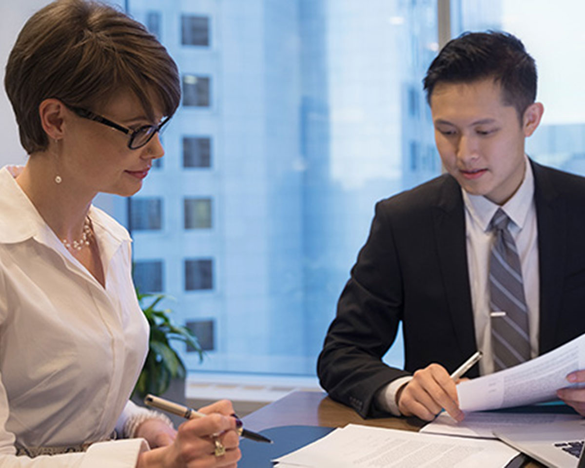 Two business professionals in formal attire reviewing and signing documents together at an office desk near a window