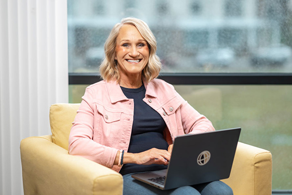 Sherry is sitting in a yellow armchair with a laptop on her lap. She’s smiling and wearing a pink denim jacket.