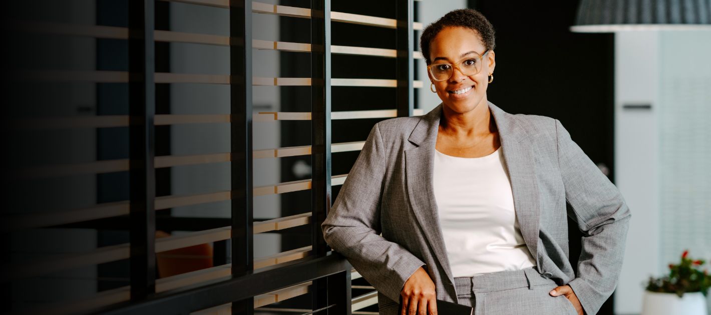 Professional wearing glasses, gray blazer and white top smiles warmly in modern office setting with horizontal window blinds