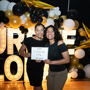 two Purdue Global graduates holding a Congrats sign, with a light-up Purdue Global sign and black and gold balloons in the background