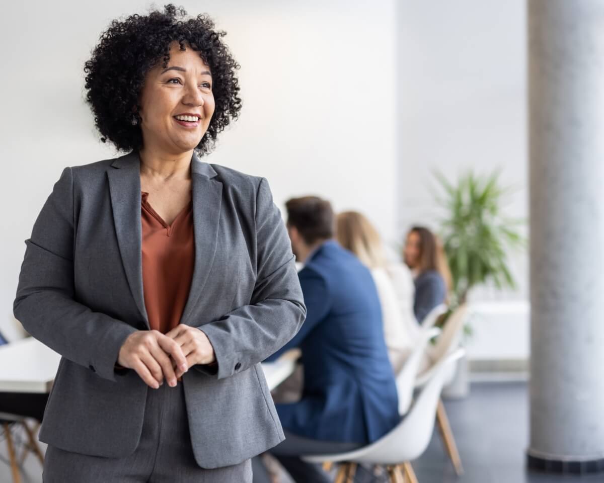A smiling businesswoman in a suit stands in an office, with colleagues at a table in the background.