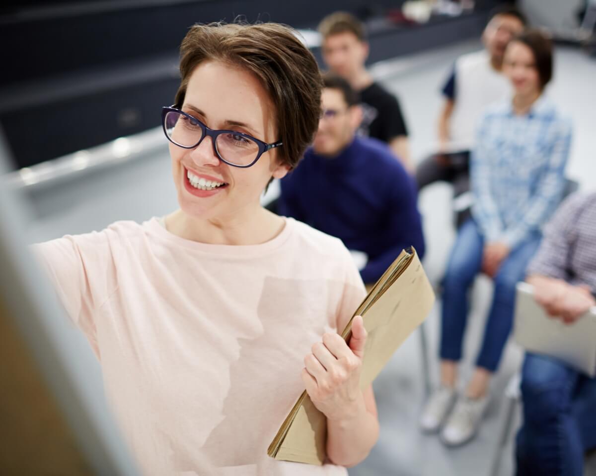 A health educator writes on a board while holding notes, with students in the background