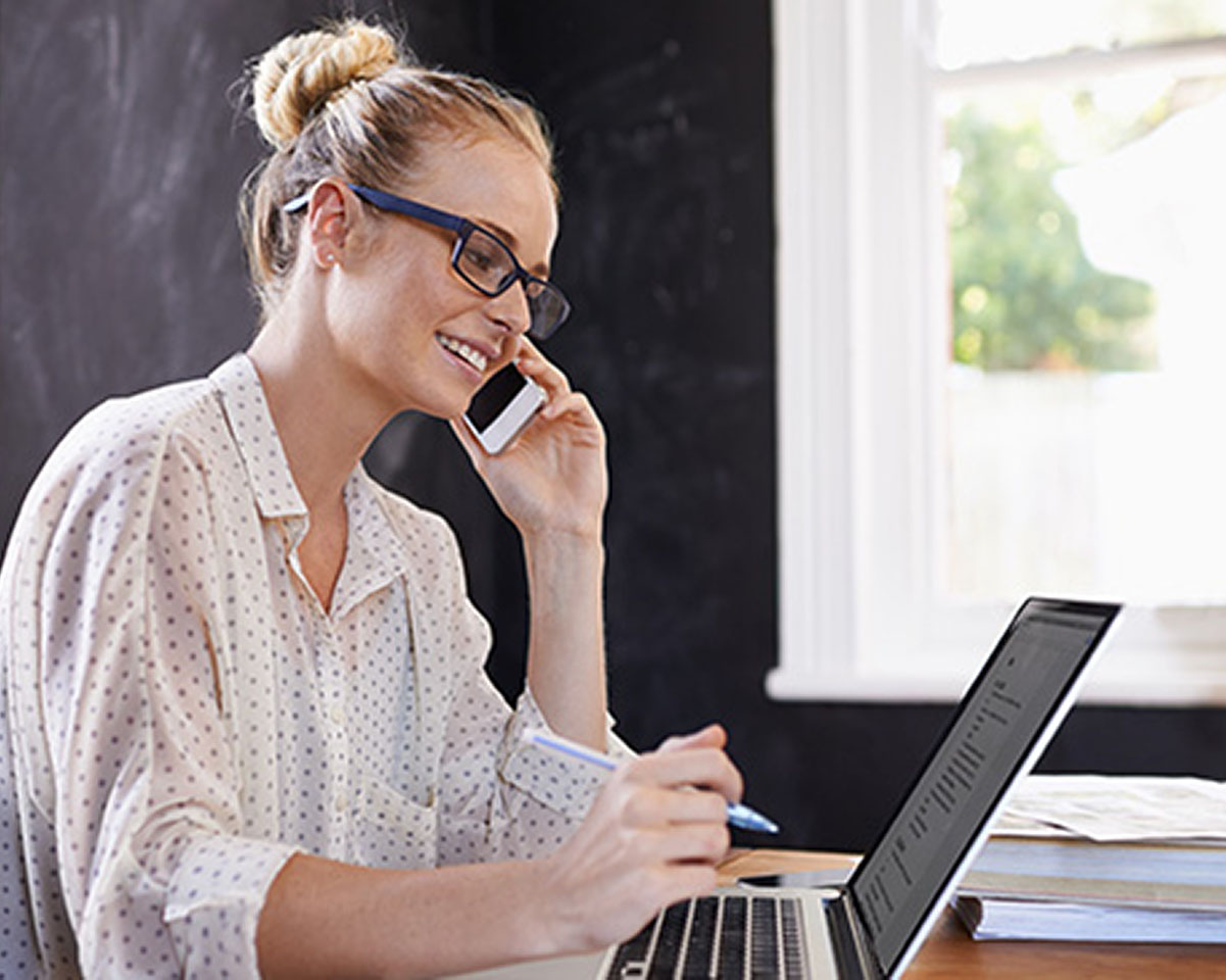 Professional in glasses and white patterned blouse smiles while working at desk with laptop and speaking on phone