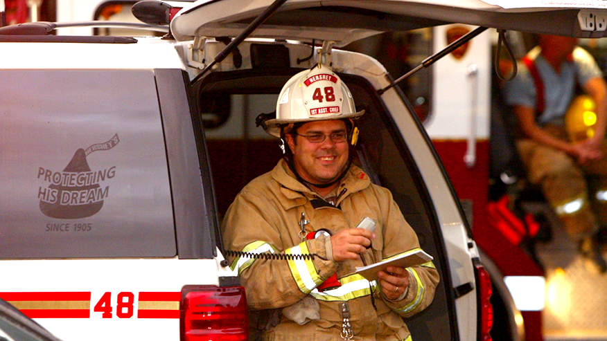 Firefighter in white hat marked with the number 48 smiles and holds a radio communicator and notebook standing near a fire department SUV.