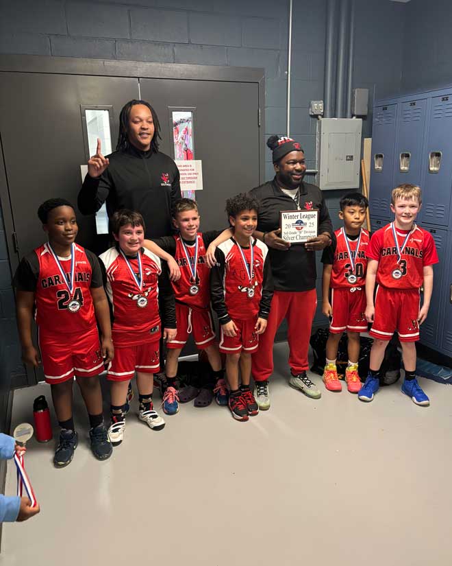 Six young boys are wearing red uniforms and posing in a line, with Gilmore and another coach (both wearing black) posing behind them.