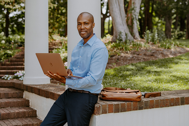 Quentin Huff leans against a brick half wall with an open laptop in hand