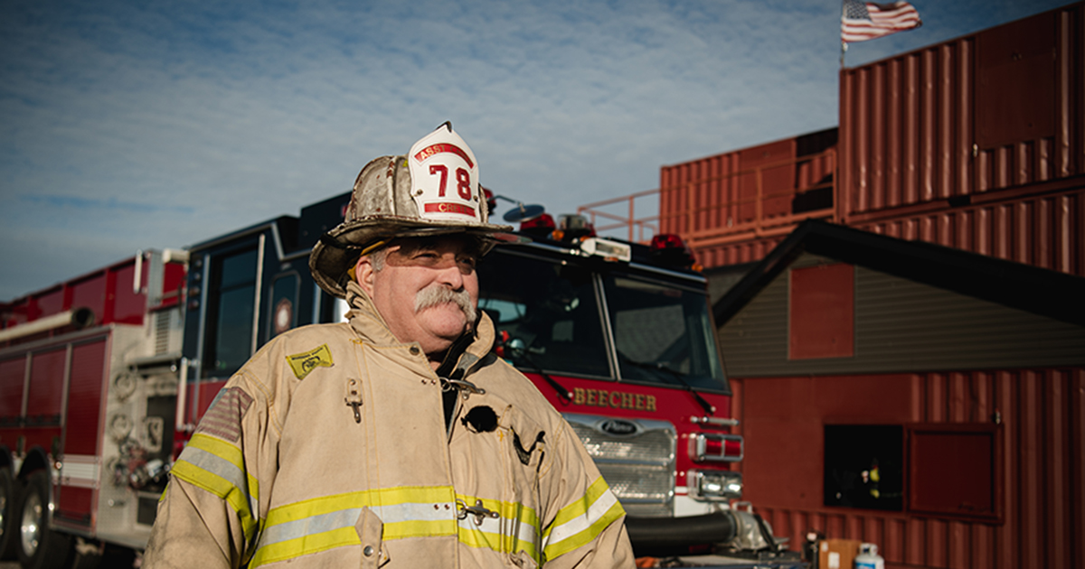 Firefighter Jonathan Mead in a fireman's uniform standing in front of a firetruck and warehouse-type building.