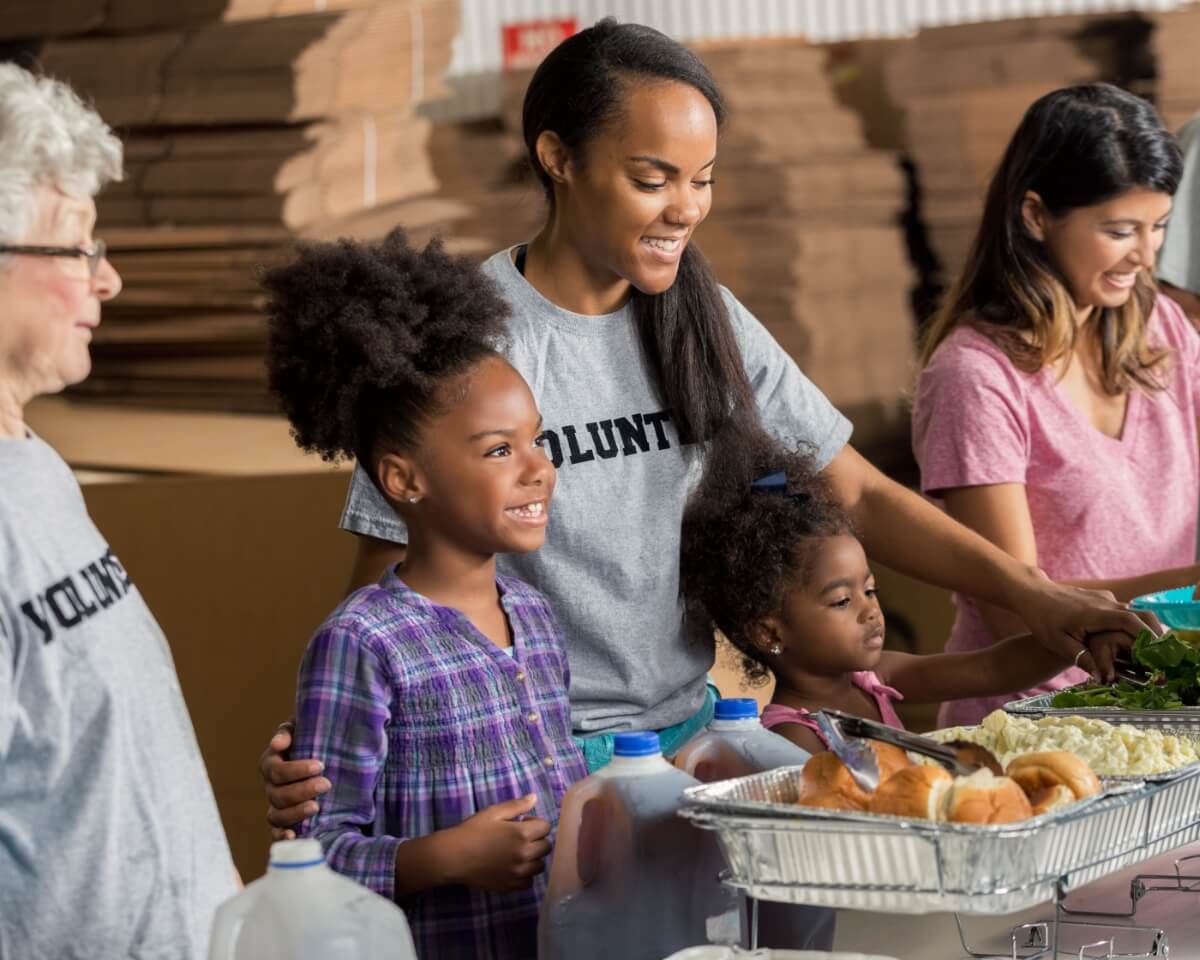 Women wearing volunteer shirts help children fill plates with food from serving pans