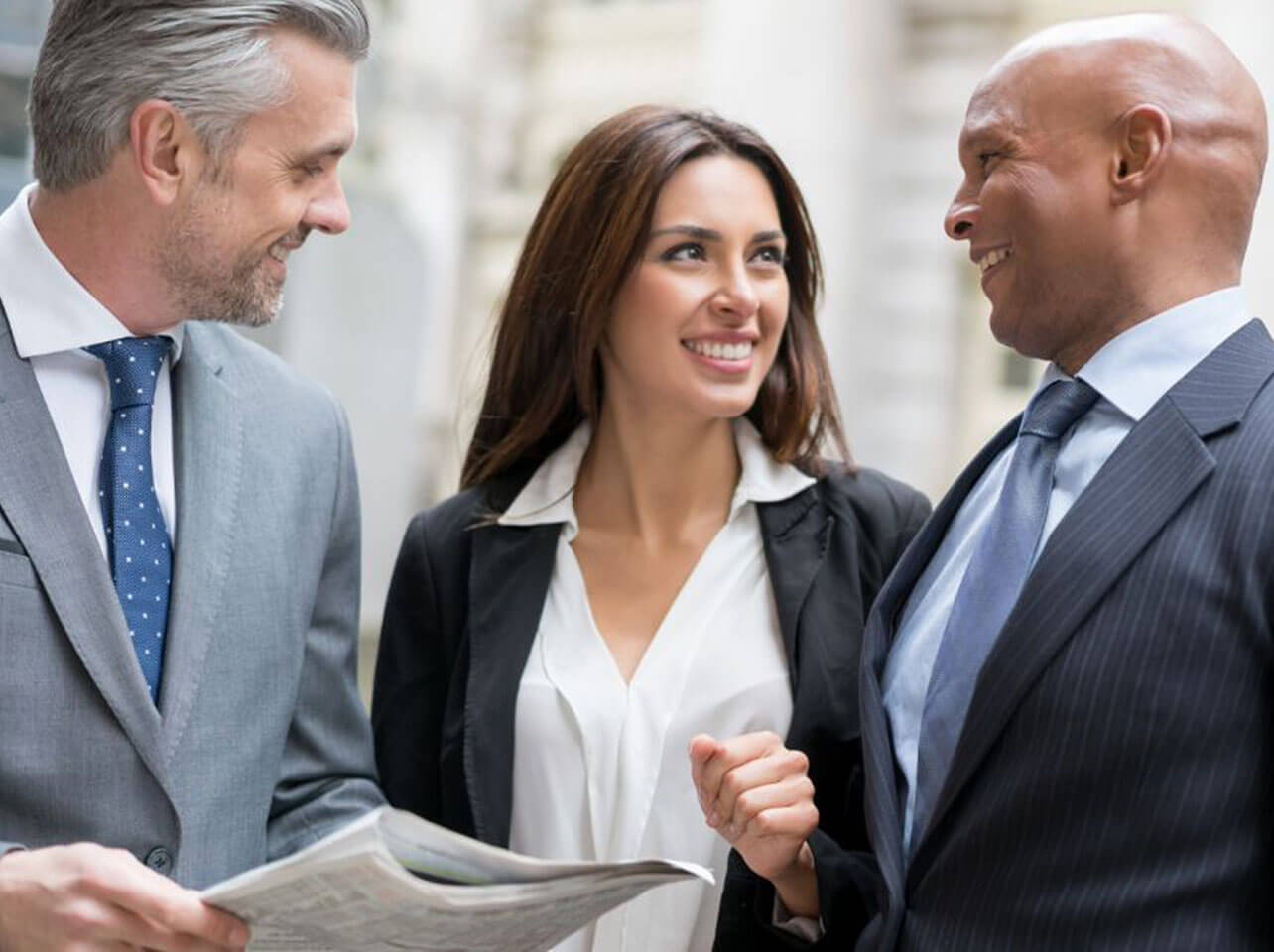 Three smiling business professionals in suits review documents together while engaged in conversation