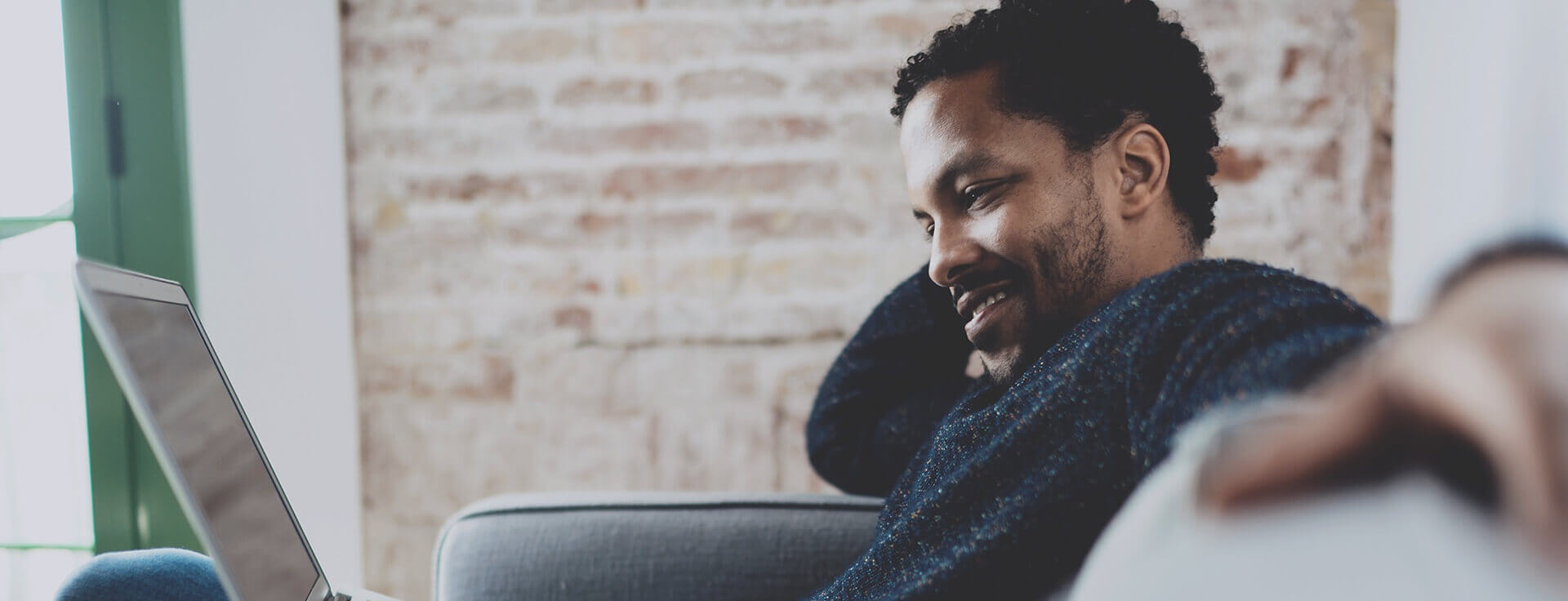Professional in a blue sweater smiles while working on laptop in modern office with exposed brick wall background