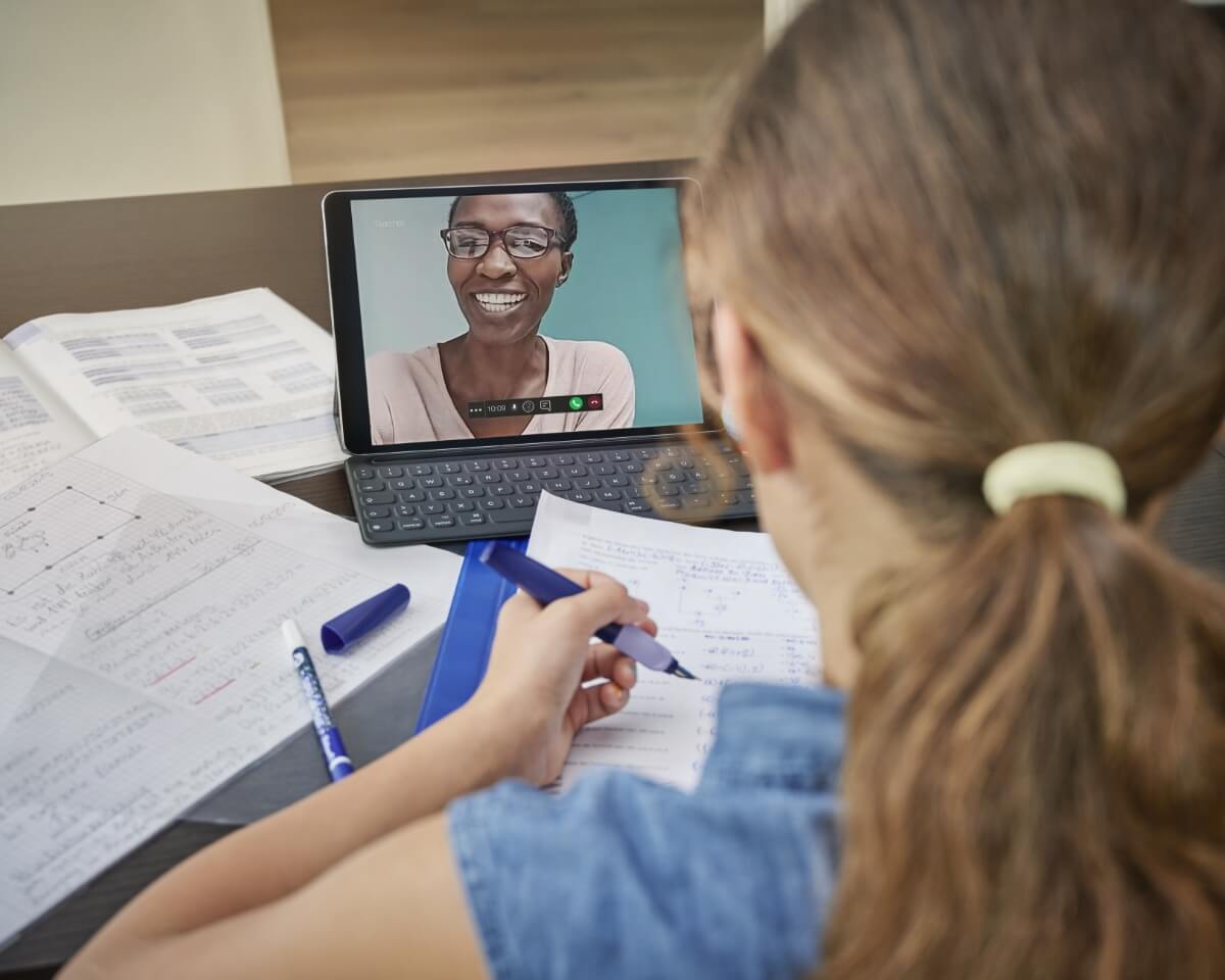woman taking notes at a desk with a laptop open and a professor visible on the screen