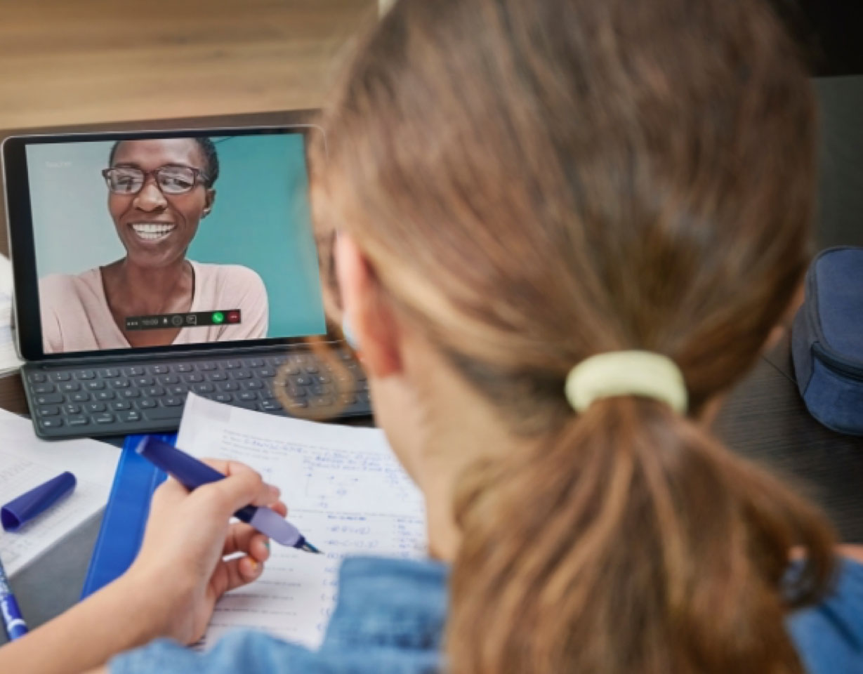 woman with her back to the camera working at a desk with a laptop open and a professor visible on the screen
