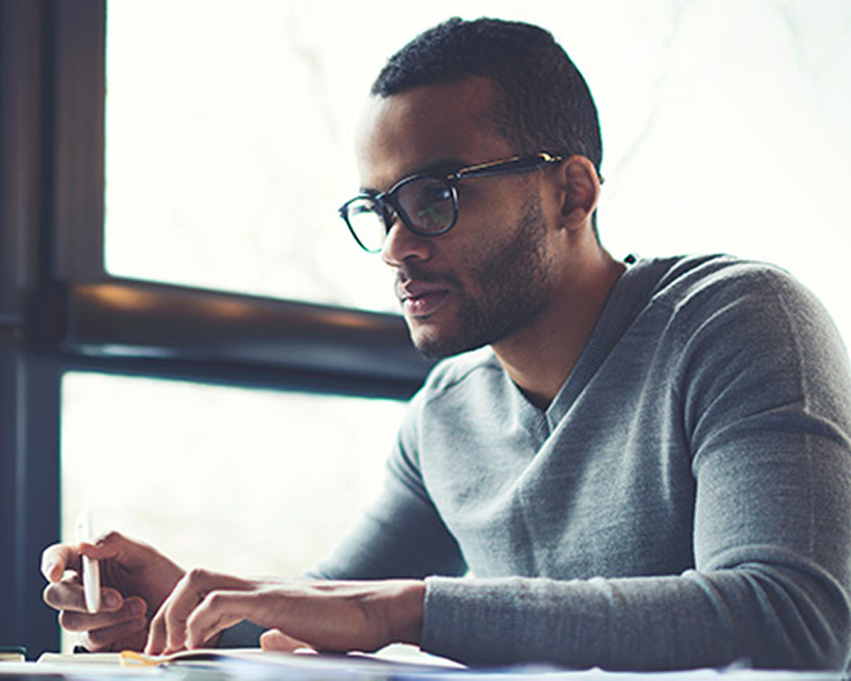 Professional in glasses and gray sweater working at desk near window, focused on documents in natural lighting