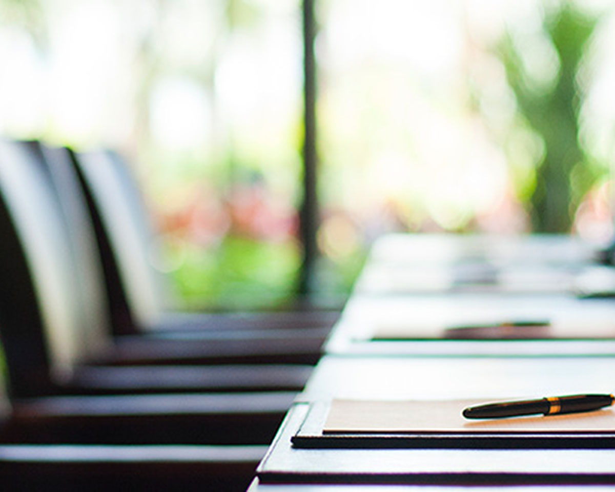 Empty conference room table with papers and pens laid out and view of blurred green background through windows