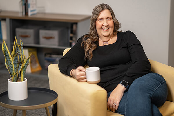 Marcy sits in a chair and smiles while drinking a cup of coffee.