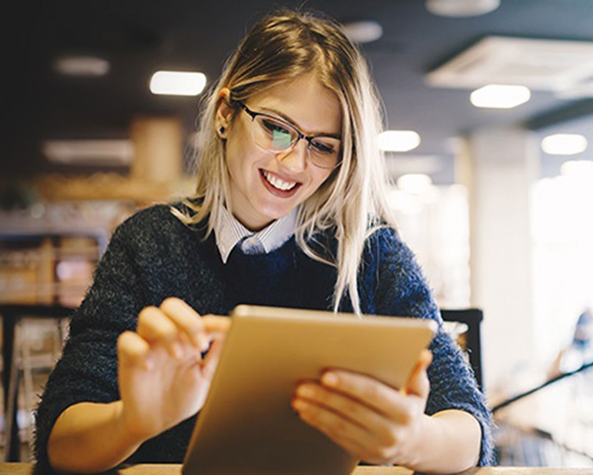 Professional in glasses and navy sweater smiles while working on tablet device in bright office setting