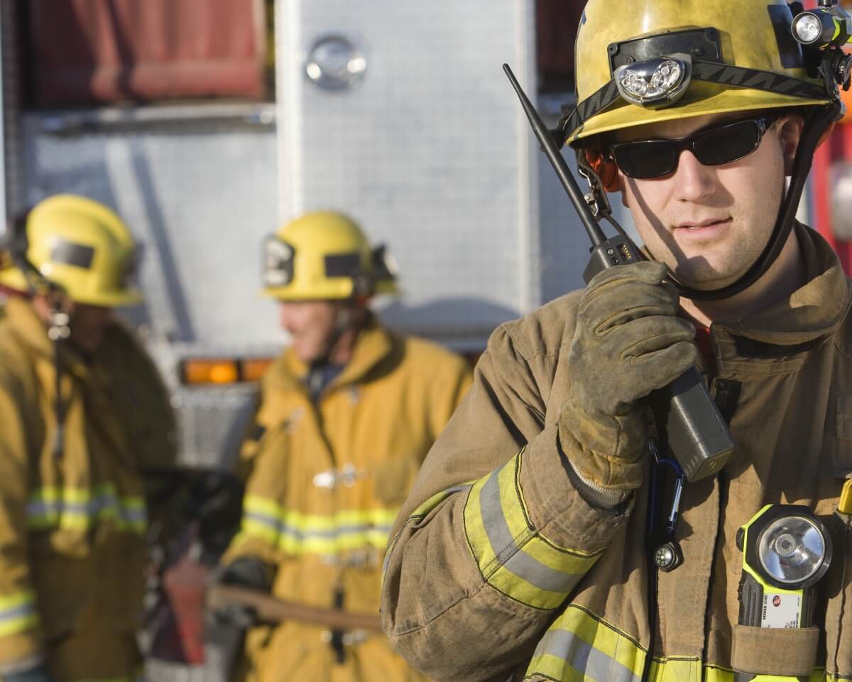 Firefighter in protective gear holds radio, with fellow firefighters in background in uniform