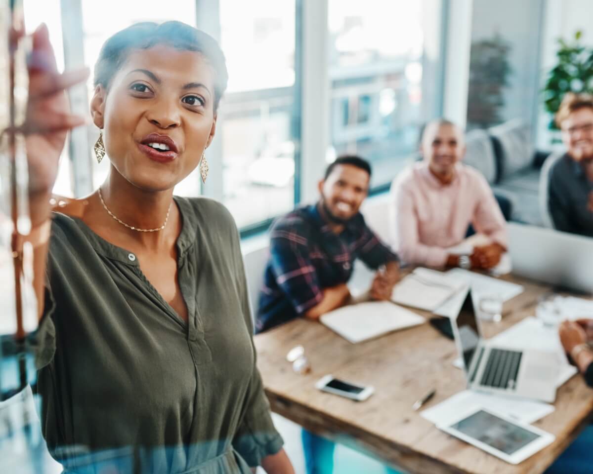 Marketing professional presents to colleagues at wooden table with laptops in a bright office.