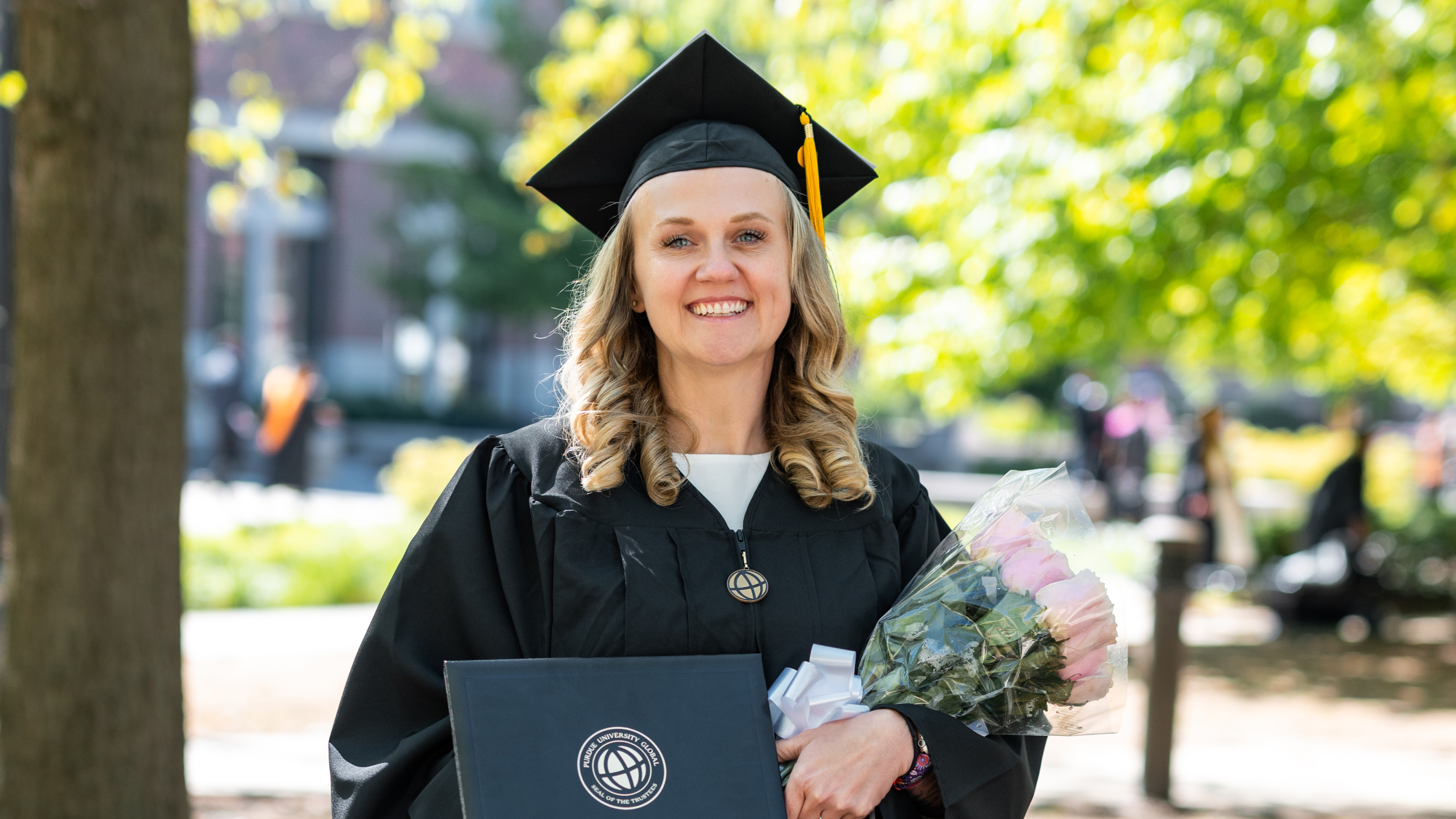 cap: Angel Riley completed Purdue Global’s RN-to-BSN program and is already moving into a new role in her career. (Purdue Global photo/Kelsey Lefever)
alt: Angel Riley is wearing a black cap and gown, holding a diploma and a dozen pink roses.