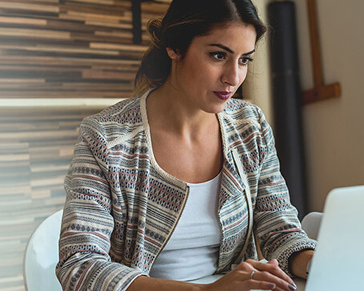 Woman in patterned jacket and white top working at laptop near window with venetian blinds casting striped shadows