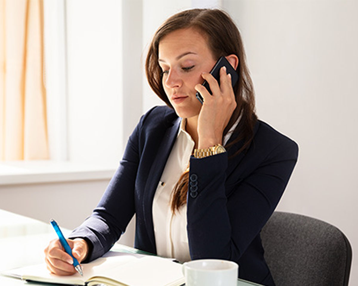 Professional in navy blazer taking notes while speaking on phone in bright office setting