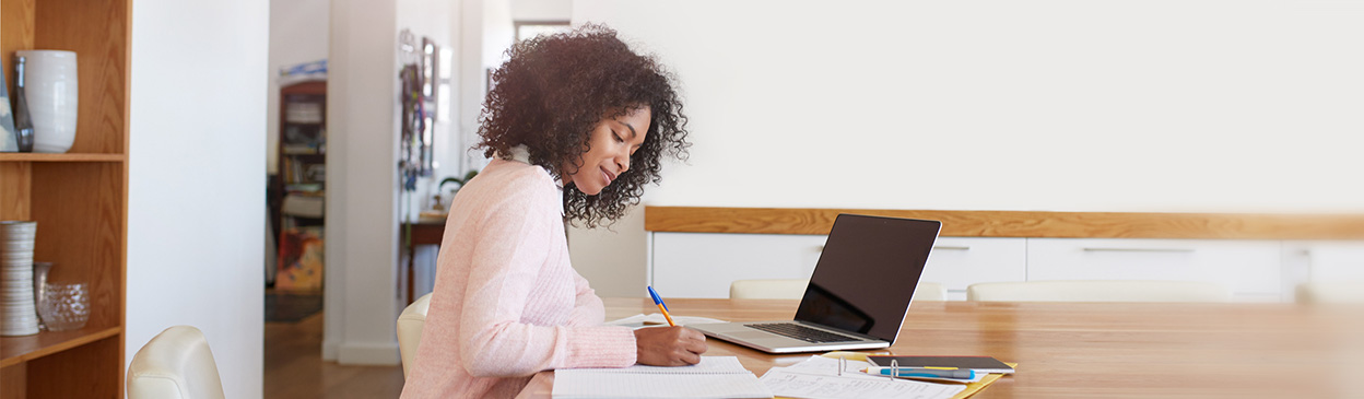 Person with curly hair wearing pink sweater working at wooden desk with laptop and papers in bright home office setting