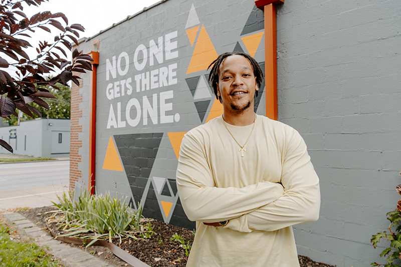 Theo Gilmore standing in front of a mural on the side of a building that says "No one get there alone."