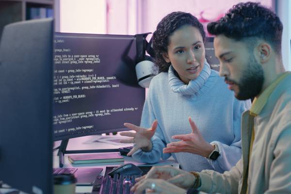 A female IT specialist and a male coder discuss ethical software development as he works at his computer.